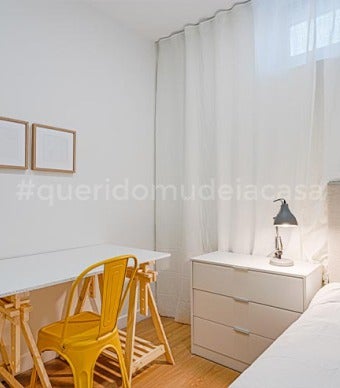 quarto de solteiro com uma secretária branca e uma cadeira amarela torrado , quadro de madeira e cortinado cinza claro e uma mesa de cabeceira com um candeeiro preto ligado